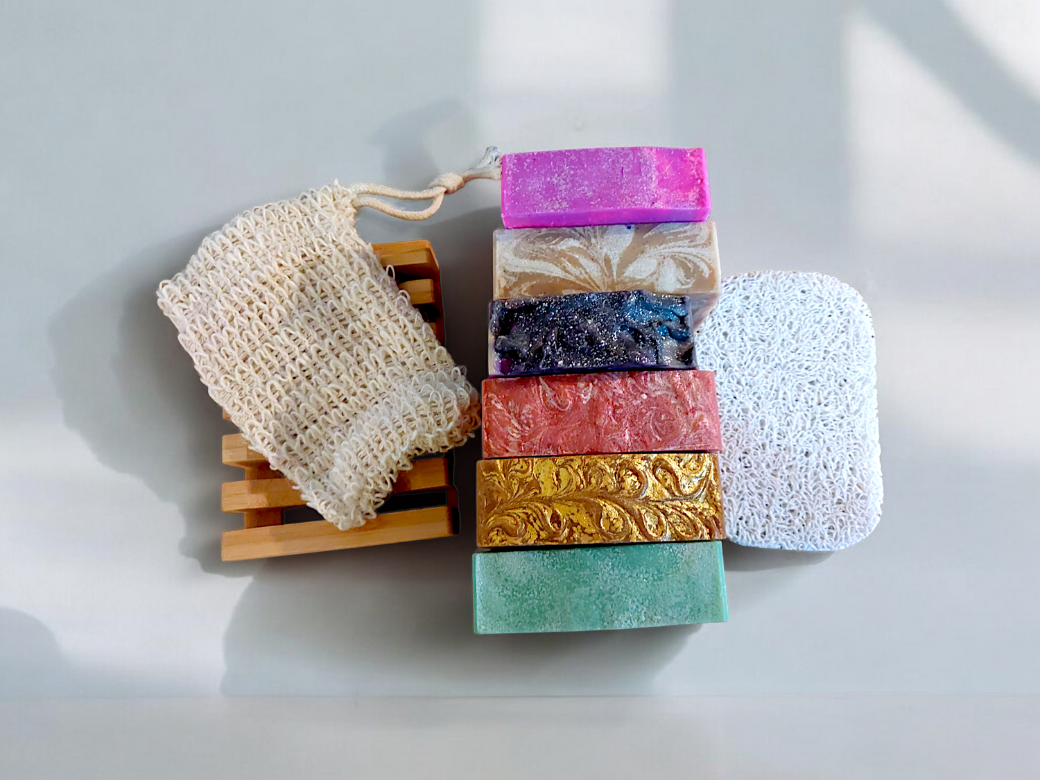 A group of six bars of colored soaps with a mesh bag and a bamboo soap tray on the left and a white soap lift on the right. 